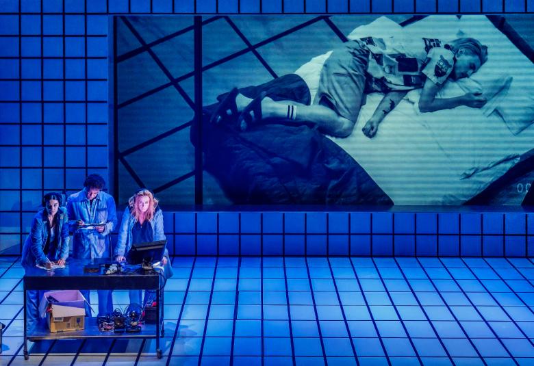 Kuvassa on sininen sävy ja alhaalla vasemmalla on kolme henkilöä pöydän ääressä. Koko huone on lattioilla ja seinillä ktasaruutuista klinkkeriä.  Seinällä on iso ruutu, josta näkyy nuori nainen makaamassa sängyssä.