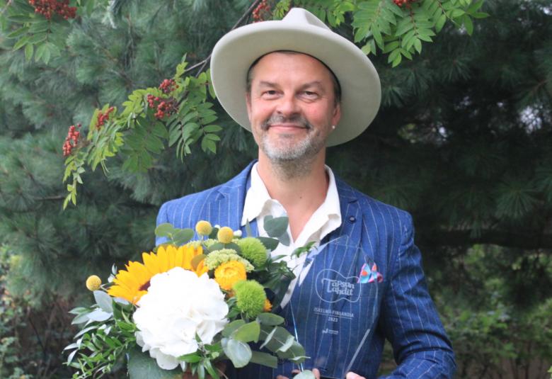 Kuvassa on Tuure Kilpeläinen puolivartalokuvassa ja hänellä on kädessään onnittelukukkaset. Taustalla on pihlajia.