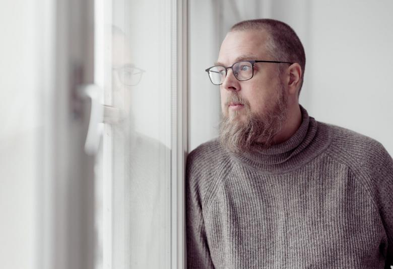 Kuvassa on Tuomas Kyrö vaaleasävyisessä huoneessa ja hän katsoo ikkunasta ulos.Hänellä on harmaa villapaita päällään.