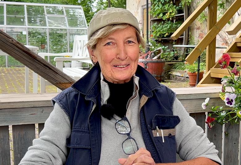 Kuvassa on Pirjo Honkasalo istumassa terassilla ja taustalla näkyy kasvihuone. Hänellä on harmaa pusero ja tumman sininen liivi päällään sekä vaalean vihreä hattu.