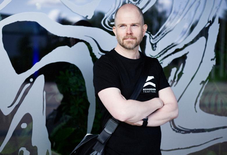 Kuvassa on Riku Innamaa puolivartalokuvassa ja hänellä on musta t-paita päällään. Kädet ovat ristikkäin edessään. Taustalla on lainehtivia musta-sini-harmaa kuvioita koko seinän laajuisesti.