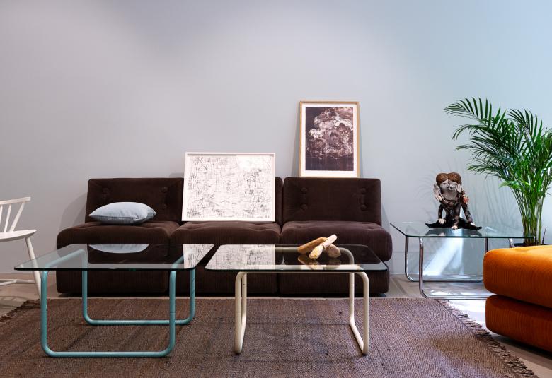 Kuvassa on olohuoneen kalustusta ja takana ruskea sohva, jonka edessä on graafinen teos.  Sohvan edessä on lasinen pöytä ja vieressä oranssi nojatuoli. Vasemmalla on valkoinen puinen tuoli.