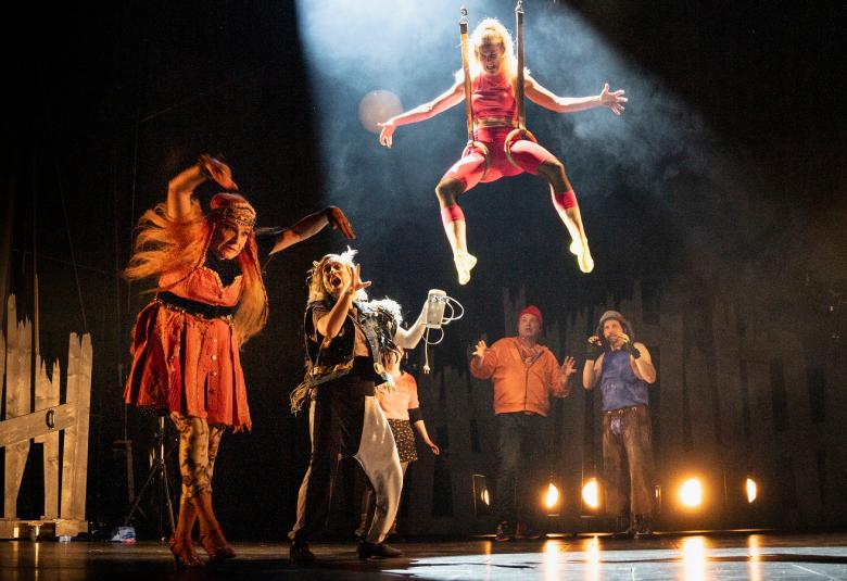 Kuvassa on näyttämöllä osavalaistuksessa ja vasemmalla on oranssissa vaatteessa ja vieressä on ringissä useita henkilöitä kalevalaisissa mystisissä asuissa.  Keskellä on ylhäällä roikkumassa nainen kädet levitettynä.