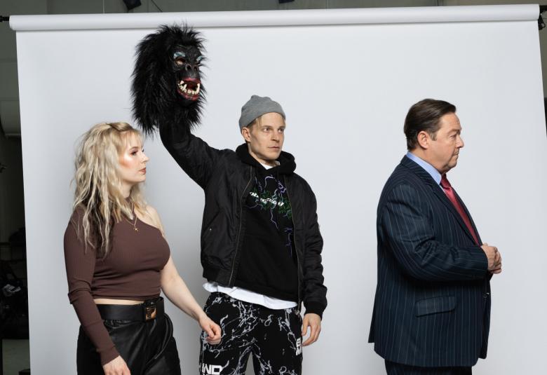 Kuvassa ovat ruskeassa puserossa ja mustissa farkuissa Anna-Sofia Tuominen, musta apinanaamari vasemmassa kädessään ylhäällä Olli Riipinen ja musta puku päällään Jouko Klemettilä poistumassa oikealle.  Taustalla on iso valkokangas.