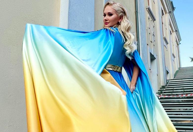 Kuvassa on Ekaterina Buzhanska Ukrainan lipun väreissä olevassa laajassa, ilmavassa mekossa. Kuva on otettu ulkona rakennuksen vieressä, jossa on portaat.