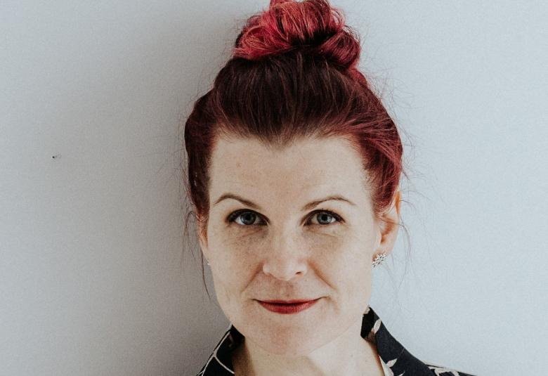 Kuvassa on Elina Pohjola kasvokuvassa.  Hänellä on punaiset hiukset ja ne on hutturalla ylhäällä. Hänellä on musta-valkoinen pusero ja avonainen kaula-aukko.