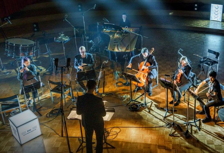 Kuvassa on Ensemble for New Music Tallinn lavalla esiintymässä.  Kuva on otettu ylhäältä alaspäin ja se on sinisävyinen.  Ylhäältä päin tulee valonheittäjät. Keskelle tulee oranssia valoa lattiaan.