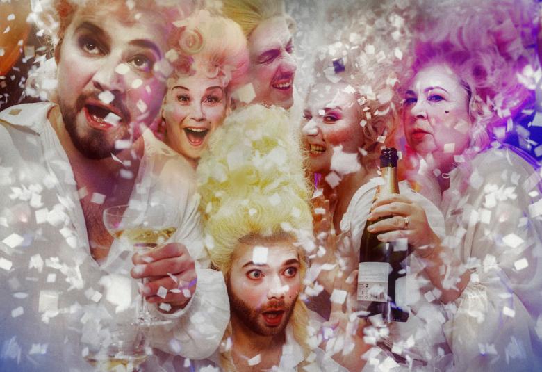Kuvassa ovat kuusi näytelmän näyttelijän kasvot kuplivassa, värikkäässä ympäristössä valkoisissa puvuissa nauttimassa sampanjaa.