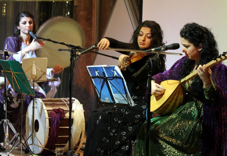 Kuvassa ovat Gûlistan Ensemblen kolme soittajaa esiintymässä ja esiintymisasut ovat tumman kiiltäviä.  Soittimina ovat rummut, viulu ja kastanja.