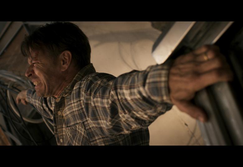 Kuvassa on Peter Franzén sivuttain roikkumassa kuilutilassa, jossa on paksuja sähköjohtoja. Kuva on puolivartalokuva ja hänellä on päällään ruudullinen paita.