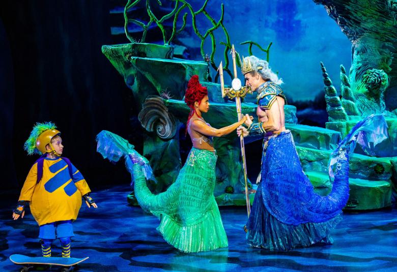 Kuvassa on keskellä vihertävässä puvussa merenneito ja oikealla on vastapäätä sinisessä puvussa kuningashahmo. Vasemmalla on skeittilaudan vieressä pikku poika kelta-sinisessä paidassa.  Valaistus on sininen.