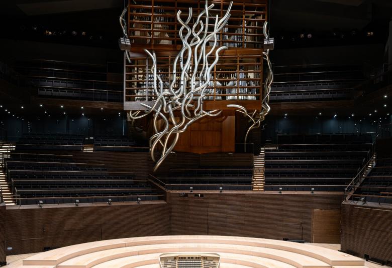 Kuvassa on Musiikkitalon konserttisali tyhjänä ja keskellä ylhäällä on teräksinen kiehkurainen iso koriste. Alla on puinen orkesterialue.  Takana on tumma katsomo ja keskellä kuva ruskeistä uruista.