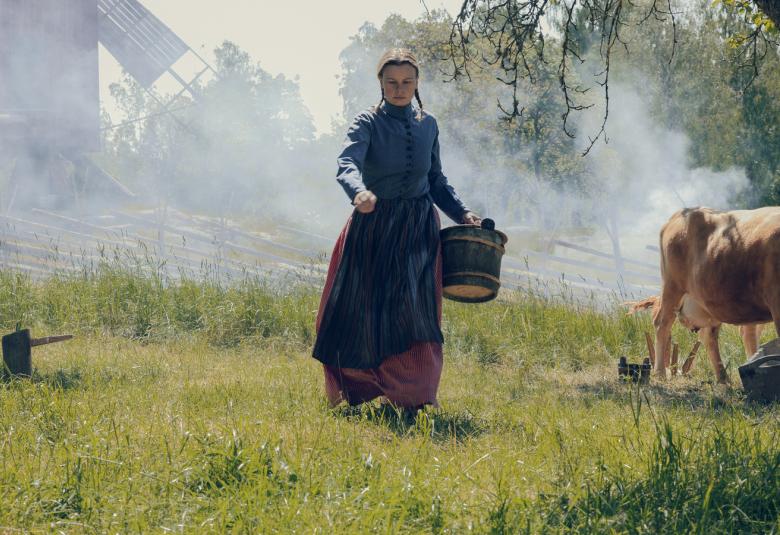 Kuvassa on Amanda Andersson kävelemässä nurmella ja taustalla on lehmisavua sekä lehmä oikealla. Vasemmalla takana tuulimylly.