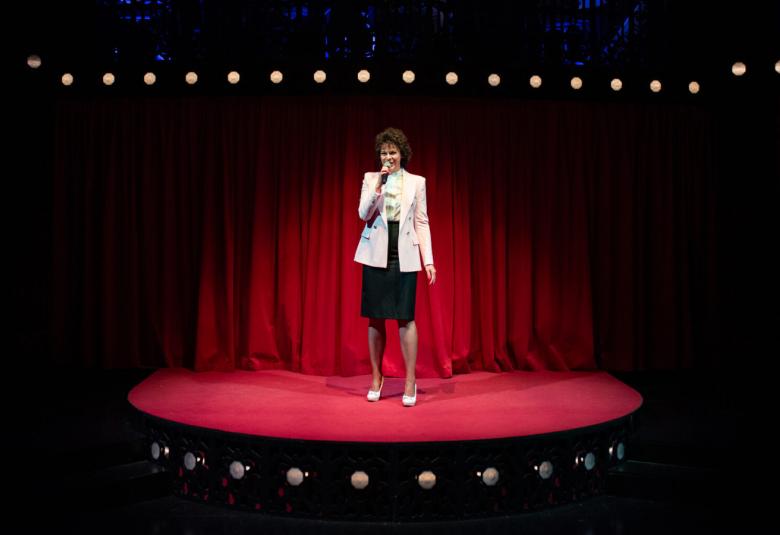 Kuvassa on Paula Koivuniemeä esittävä Ninni Kekki lavalla ja hänellä on päällään valkoinen jakku ja musta puolihame.  NÄyttämön valokeilassa näkyy punainen lattia ja verhot.