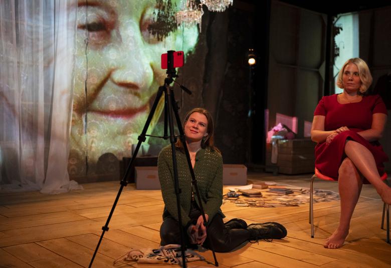 Kuvassa ovat Inke Koskinen lattialla ja Emmi Kaislakari istumassa tuolissa. Edessä on kamera ja taustalla on heijastettuna Seela Sellan kasvot.