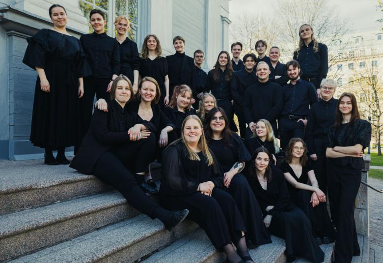 Kuvassa ovat kuorolaiset mustissa asuissa. Heistä taaimmaiset seisovat vanhan valkoisen rakennuksen edessä ja osa istuu portailla.