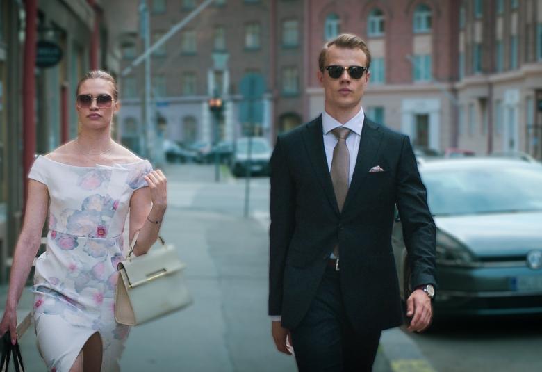 Kuvassa ovat Tuulia Eloranta vaaleassa kotelomekossa ja Samuel Kujala tummassa puvussa kävelemässä kadulla. Taustalla on autoja parkkeerattuna.