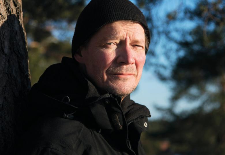 Kuvassa on Juha Hurme tumma pipo päässään.  Hänestä näkyy kasvot. Vasemmalla on männyn runkoa ja oikealta tulee valoa. Muu osa on tummaa.