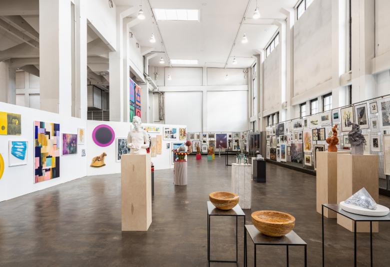 Kuvassa on näkymä näyttelyhallista jossa on etualalla valkoisten jalustojen päällä puuesineitä.  Molemmin puolin ja takana ovat seinämät, joilla on graafisia teoksia. Seinät ovat valkoisia.