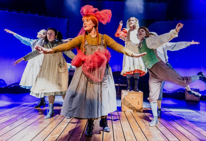 Kuvassa on tyttöjen mekoissa neljä näyttelijää ja yksi poika housuissa ja paidassa tanssimassa. Edessä on Anna isossa punaisessa rusetissa ja harmaassa mekossa. Tausta on kirkkaan sininen.