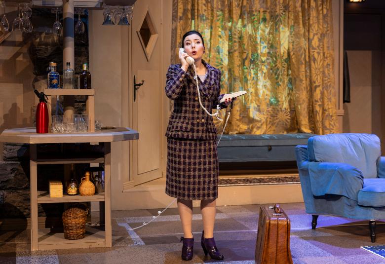 Kuvassa on Anna-Maija Oka seisomassa keskellä olohuonetta, jossa näkyy oikealla vaalean sininen nojatuoli.  Oka seisoo keskellä huonetta ruskearuudullisessa jakkupuvussa.