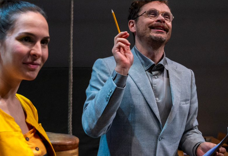 Kuvassa on Jussi Lehtonen opettajana harmaassa takissa ja osoittaa ylöspäin kynä kädessään.  Vasemmalla on Denisa Snyder keltaisessa puserossa  ja katsoo vinosti oikealle.