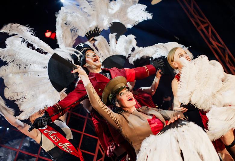 Kuvassa on keskellä takana miesnäyttelijä koppalalkissa ja punaisessa takissa sekä valkoisia höyheniä takanaan.  Edessä keskellä on kaksi näistä, joista vasemmalla olevalla on ruskea sotilastakki ja oikealla olevan edessä on valkoisea höyheniä.