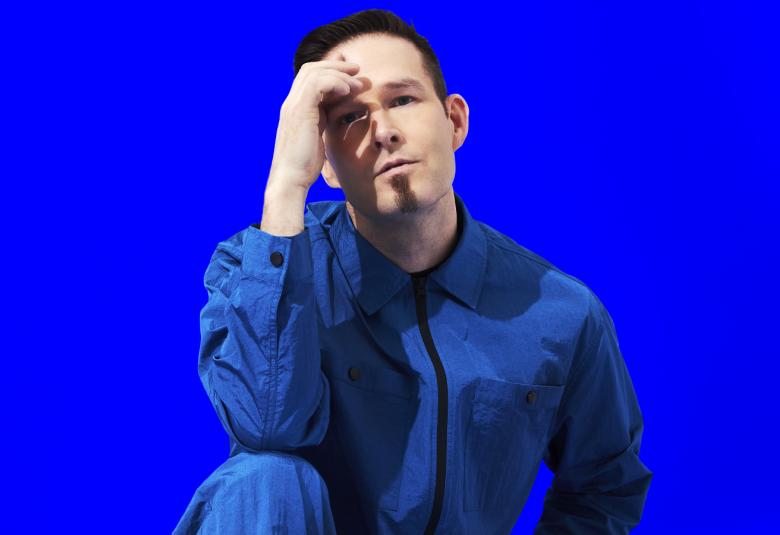 Kuvassa on Darude polviasemmossa ja nojaa oikealla kädellään oikeaan polveensa. Hänellä on siniharmaa farkkutakki ja tausta on sähkönsininen.