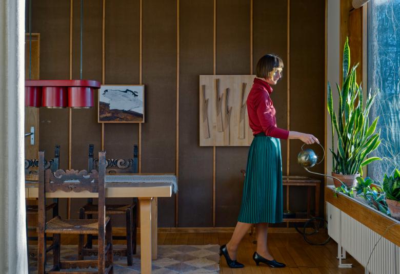 Kuvassa on huone, jossa on ruskeaa seinää ja oikealla on vaalea pöytä ja ruskea tuoli. Oikealla on ikkunaseinä ja ikkunan edessä seisoo sivuttain nainen. Hänellä sininen puolihame ja punainen pusero.