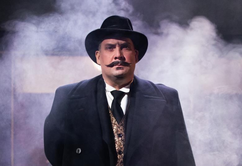 Kuvassa on Ville Majamaa Hercule Poirotina ja hänellä on päässään knallimainen lierihattu.  Taustalla on vaaleata sumua ja muuten tummaa. Majamaalla on tumma päällystakki päällään.