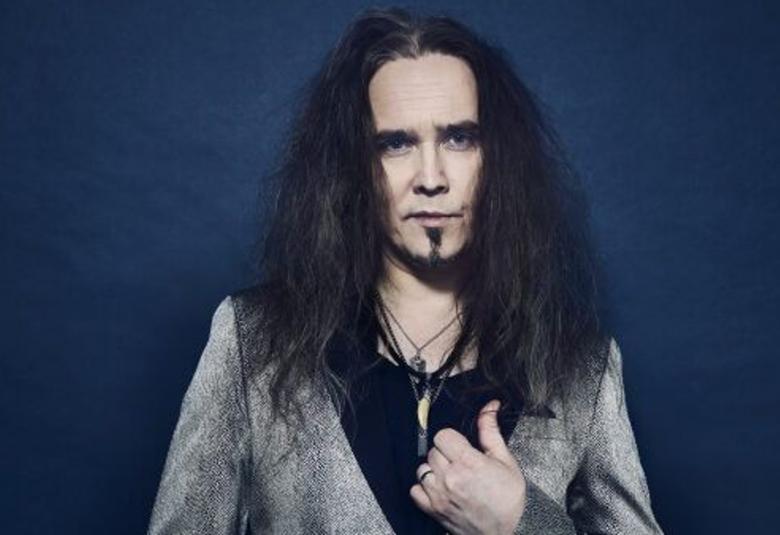 Kuvassa on Jarkko Ahola seisomassa ja hänellä on harmaa takki ja musta t-paita.  Tausta on tumman sininen.