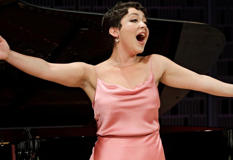 Kuvassa on Juliana Zara puolivartalokuvassa ja hänellä on kädet ojennettuina molempiin suuntiin.  Hänellä on roosan värinen kiiltävä hihaton puku.