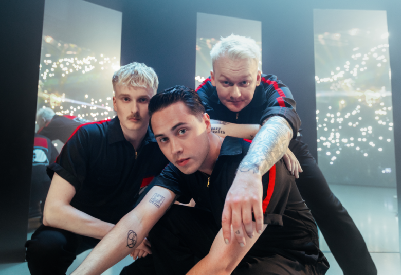 Kuvassa ovat KUUMAA-yhtyeen kolme jäsentä istuvassa asennossa ja yksi on toisten takana.  Heillä on mustat t-paidat, joissa on punaiset raidat.  Taustalla on tumman harmaata taustaa, jossa on kolme valoikkunan muotoista ruutua.