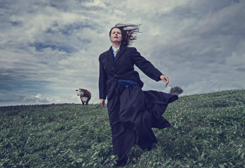 Kuvassa on Emma Klingenberg  seisomassa ruohopellolla. Hänellä on pitkä tumma hame ja takki. Taustalla näkyy valko-ruskea lehmä ja vakoisen harmaata taivasta.