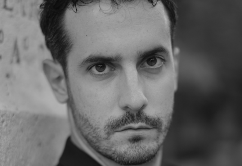 Kuvassa on Lorenzo Passerinin kasvokuva ja oikealta näkyy sementtiseinää, jossa on kirjaimet M ja A. Kuva on musta-valkoinen.