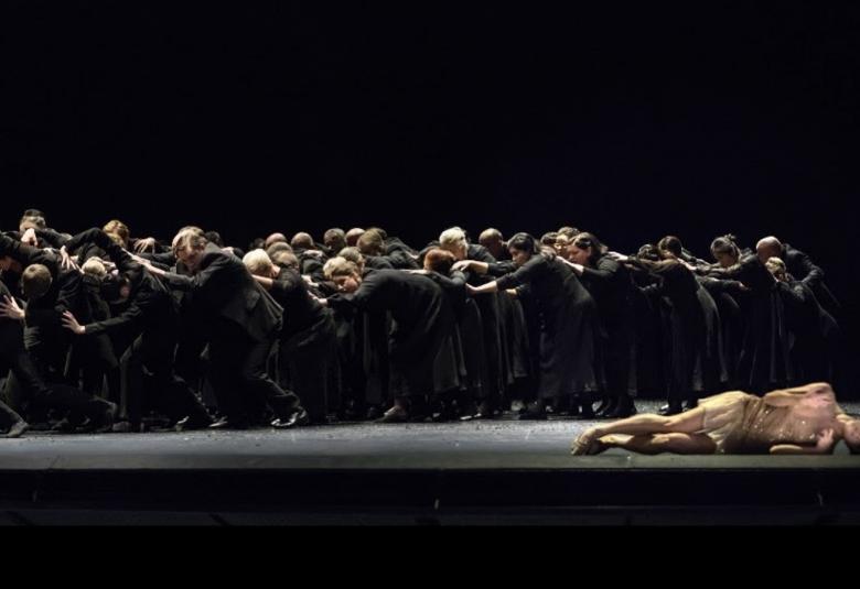 Kuvassa on yksi balettitanssija lavan lattialla oikealla ja taustalla on kumarassa vasemmalle iso joukko tanssijoita.  Kuvan sävy on tumma.