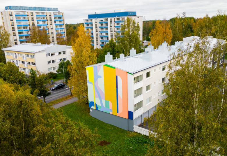 Kuvassa on ylhäältä päin kuvattu kerrostaloalue. Lähimpänä on talo, jonka päädyssä on muraali ja jossa on pystysuoria abstrakteja kuvioita eri raikkailla väreillä kuten vaaleanpunainen, oranssi ja vaalean sininen.