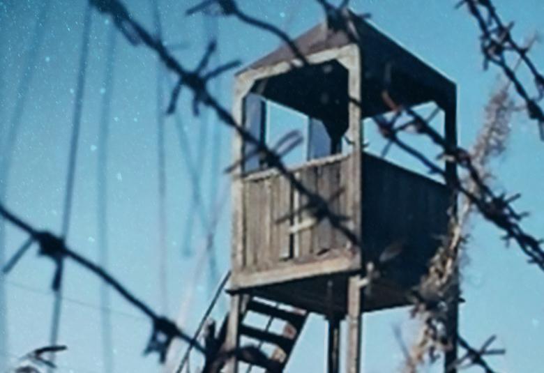Kuvassa on sinistä taivasta vasten harmaa, puinen vahtitorni. Edessä on piikkilankaa.
