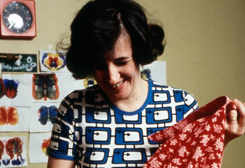 Kuvassa on nainen kumartuneena avaamaan laatikkoa, josta ottaa esiin punaisen vaatteen. Hänellä on tummat hiukset ja sinivalkoinen ruudullinen mekko päällään.