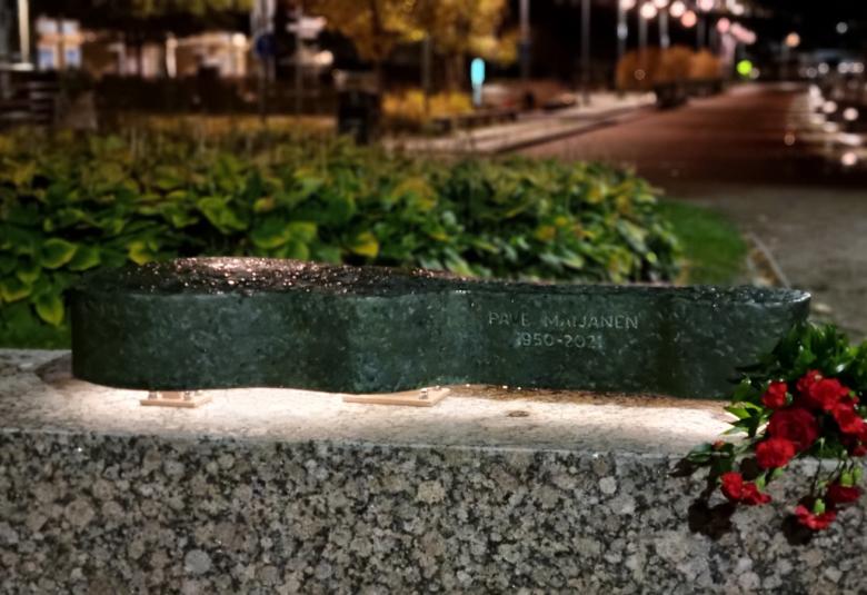 Kuvassa on kitarakotelon veistos iltavalaistuksessa. Vasemmalla paaden päällä on tumman punaisia kukkia, jotka roikkuvat osittain alaspäin.