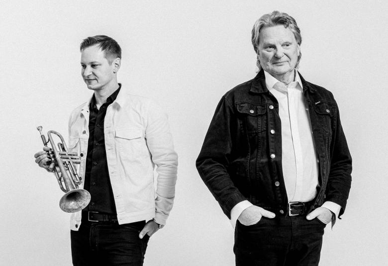 Kuvassa ovat Jukka Eskola trumpetti oikeassa kädessään ja valkoisessa farkkutakissa ja mustassa paidassa. Oikealla on Pepe Willberg mustassa farkkutakissa ja valkoisessa paidassa. Kuva on musta-valkoinen. 