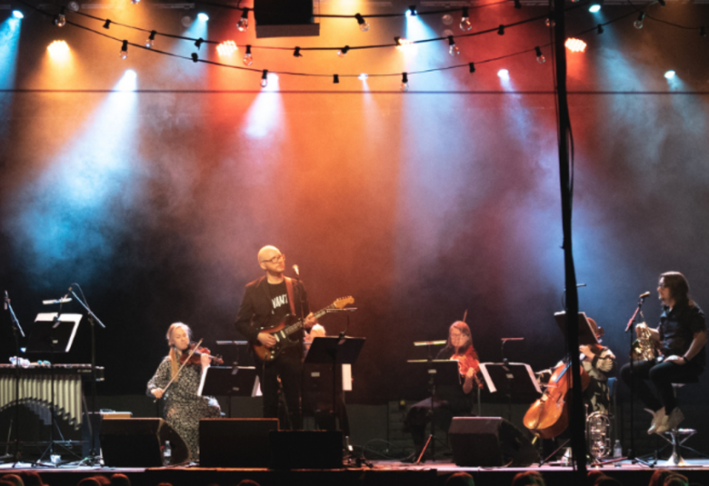 Lavalla on kansanmusiikkiyhtye, jossa on tummalla taustalla vasemmalta viulisti, kitaristi laulamassa, viulisti, sellisti ja viulisti. Taustalla on värikkäitä kohdevaloja.