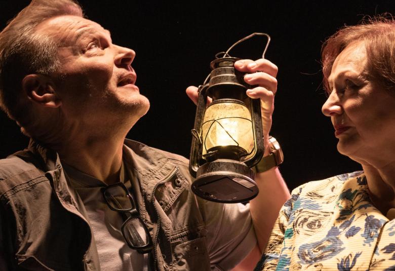 Kuvassa ovat Petri Liski ja Pirjo Luoma-aho istumassa pöydän ääressä. Liski pitää ylempänä kohotettuna vasemmalla kädessään öljylamppua. Valaistus on ruskehtava.