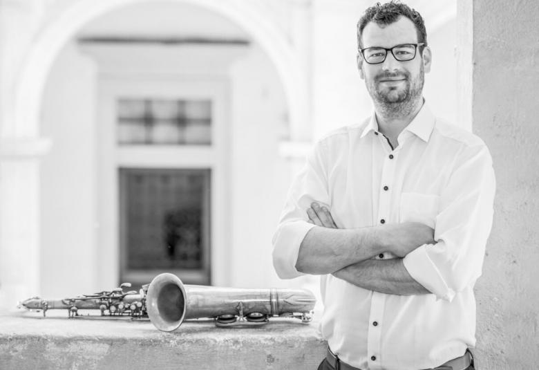 Kuvassa on Tobias HOffman ja hän seisoo kuvan oikealla puolella. Kuvan vasemmalla puolella on betonisella kaiteella saksofoni.  Taustalla näkyy valkoinen holvikaari ja taustalla ikkuna, jonka yläpuolella on kuusiruutuinen matala ikkuna.