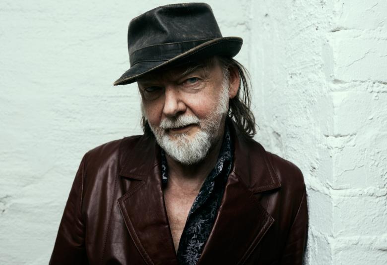 Kuvassa on Tuomari Nurmio puolivartalokuvassa ja hänellä päässään tumma lierihattu.  Katse on hieman alaviistoon ja hänellä on tumman ruskea takki päällään.  Tausta on vaalean vihertävän sininen seinä.