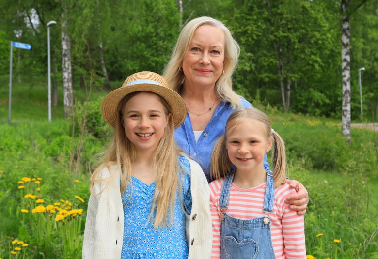 Kuvassa ovat Tiina Nopola ja hänen edessään ovat Eedla Höglund vaalean sinisessä mekossa ja Klaara Höglund vaalean sinisessä haalarissa. He seisovat vihreässä kesäympäristössä.