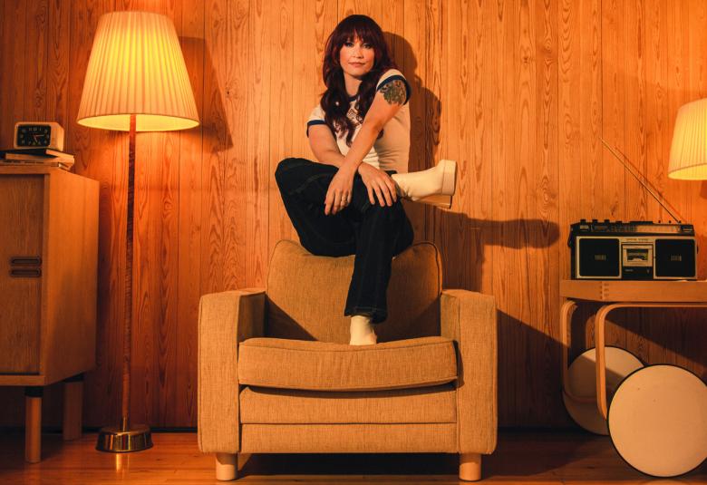 Kuvassa on Maija Vilkkumaa istumassa kellertävän nojatuolin selkänojan päällä.  Vieressä kellertävä jalkalamppu ja 1960-luvun kaappi.  Oikealla on pöytä, jonka päällä on kasettiradio. Kuvan savy on kellertävä.