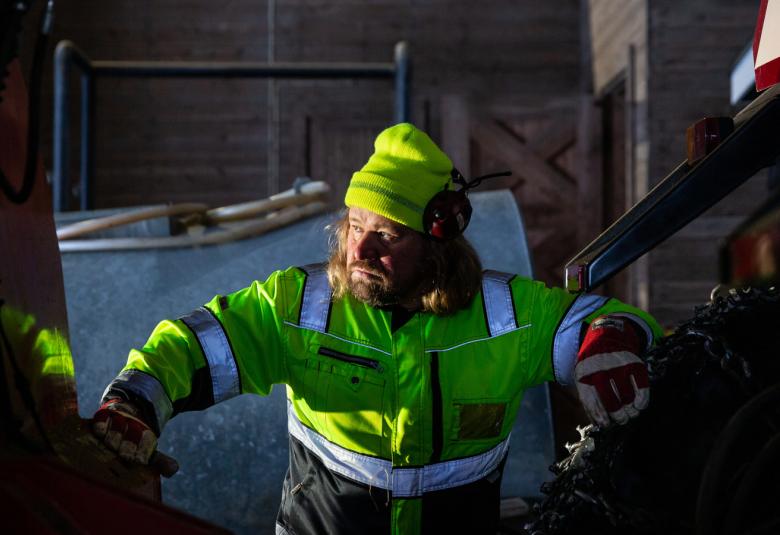 Kuvassa on Janne Kinnunen kirkkaan vaalen vihreä turvahaalari ja pipo päässään. Ympärillä on kkoneita ja latoa.