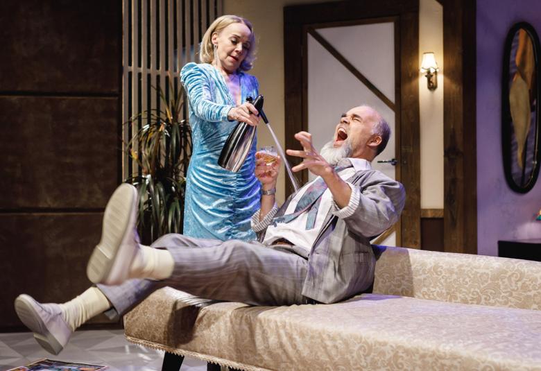 Kuvassa on Satu Taalikainen suihkuttamassa teräspullosta Veli-Matti Karénin päälle. Hän on lähes makuuasennossa sohvalla ja huutaa.  Hänellä on vaalean harmaa.