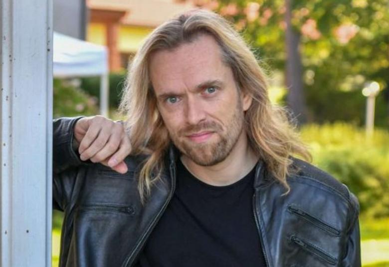 Kuvassa on Anssi Kela, joka nojaa valkoiseen kapeaan pylvääseen.  Hänellä on pitkät, vaaleat taipuisat hiukset ja hiukan partaa. Taustalta näkyy keltaista taloa ja pihaa, jossa on vaaleat teltta.